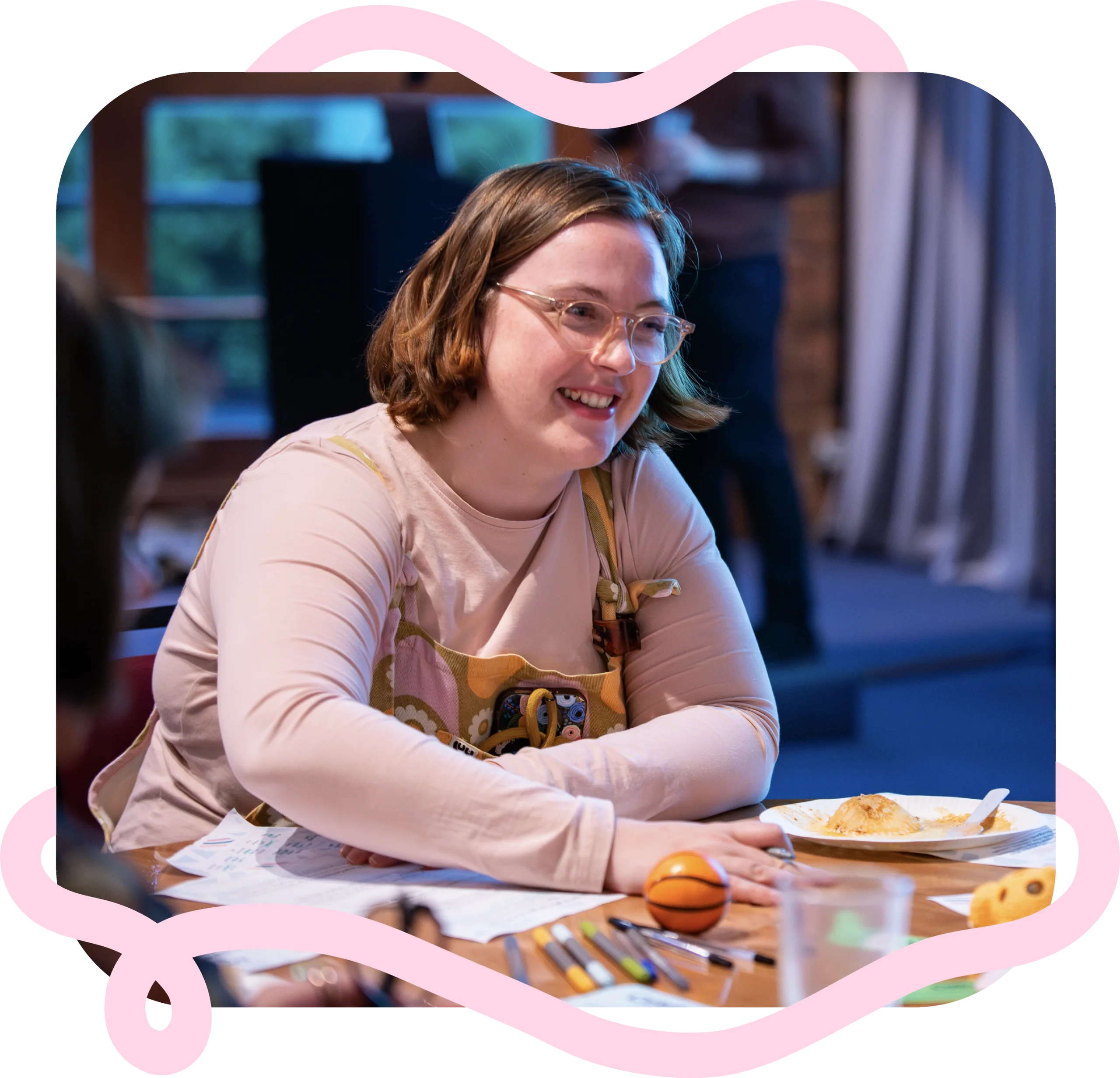 A person with glasses, shoulder length hair and dungarees sits at a table, smiling. On the table are pens, papers and some food